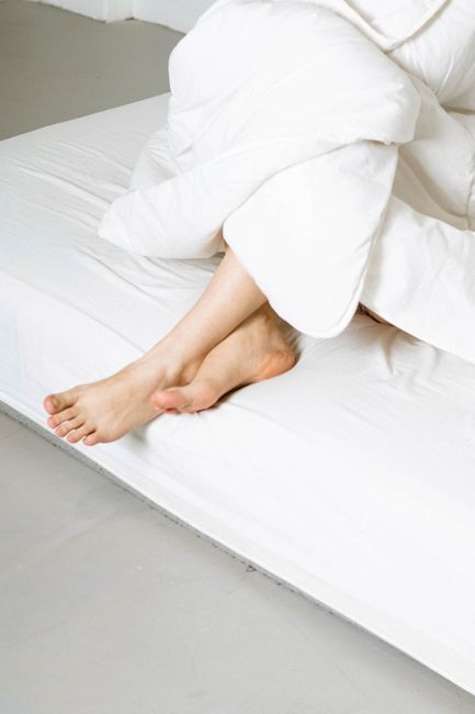 A neatly made double bed with two white pillows and a white duvet, positioned against a wooden headboard. The room features a softly lit bedside lamp on the right side, casting a warm glow. The backgr