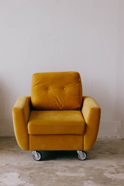 A mustard yellow upholstered armchair with a cushioned backrest and armrests, positioned on a concrete floor against a plain white wall. The armchair has visible buttons on the backrest and rests on s